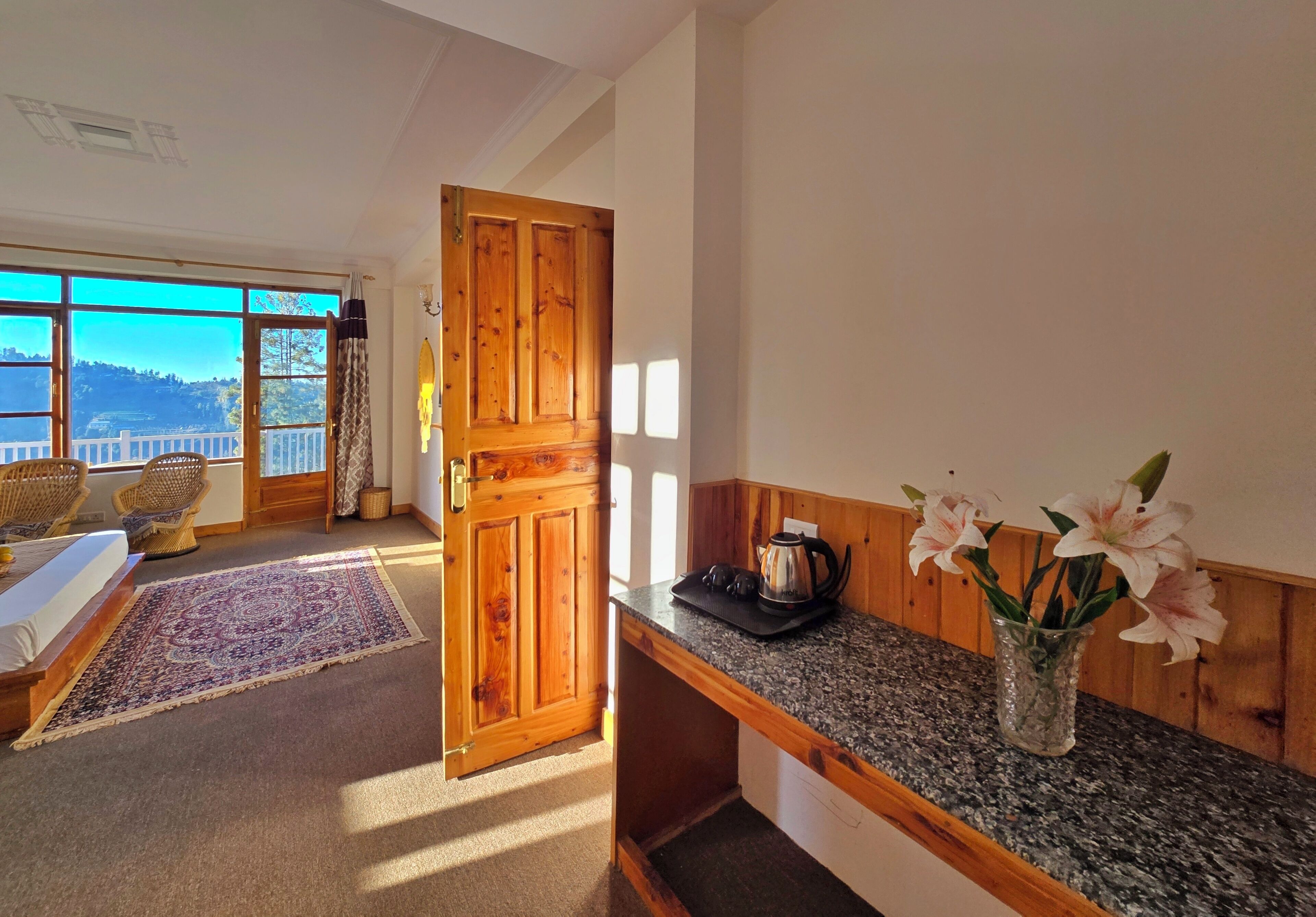 A room with a wooden door, a bed with a view, a kitchen counter with flowers, and a view of the outdoors.