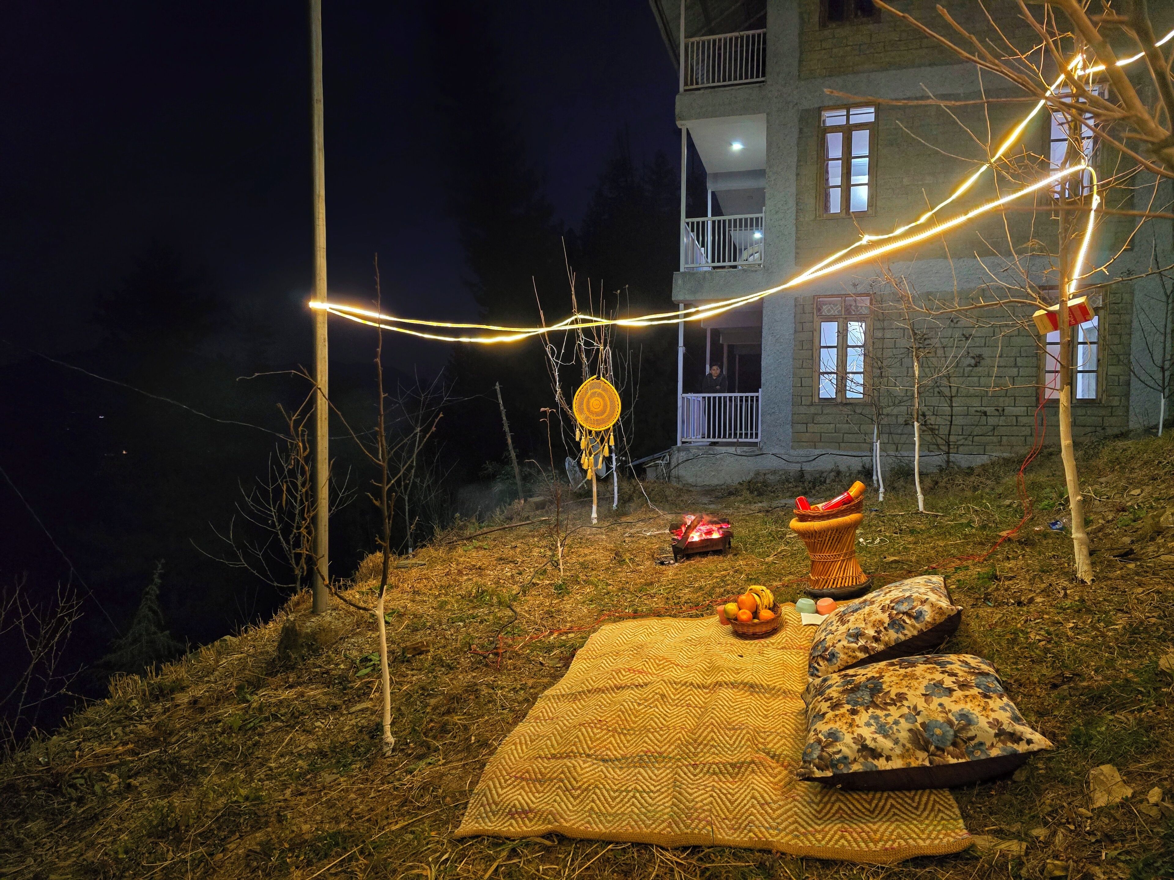 A cozy outdoor seating area with a blanket and pillows, a basket of fruit, and a small fire.