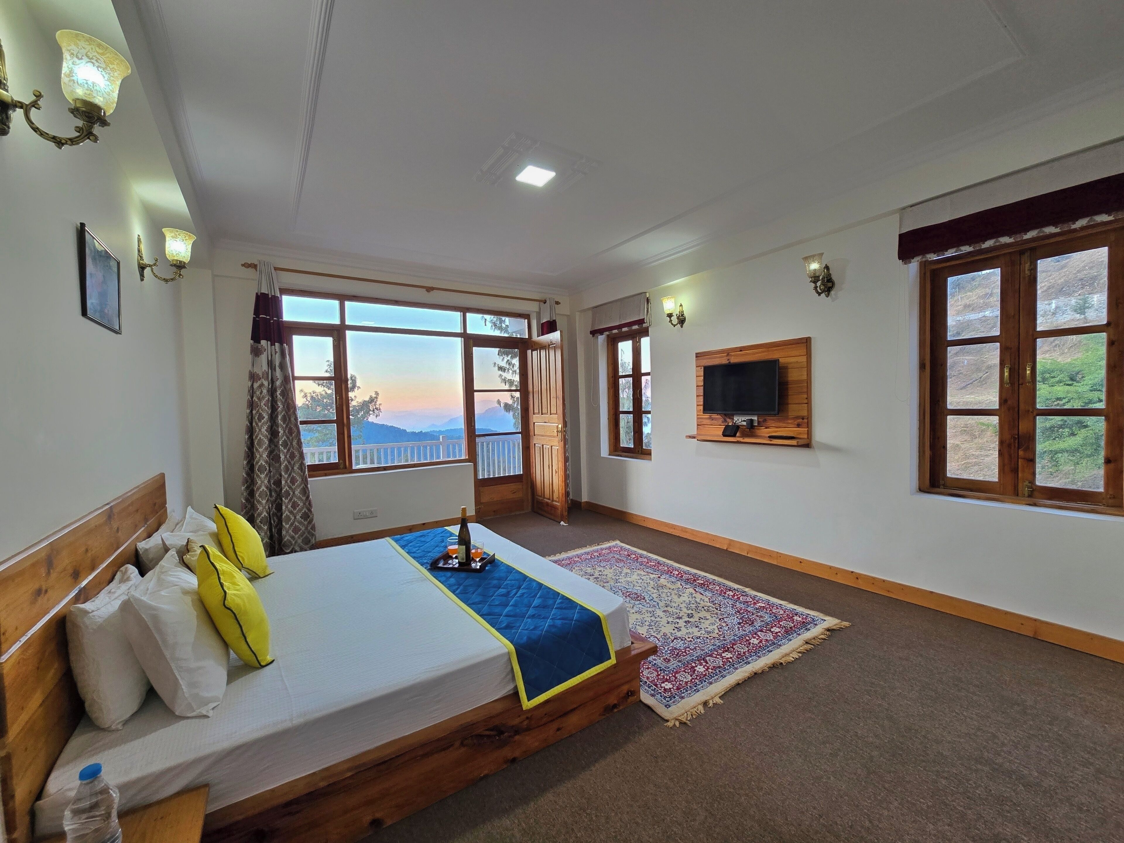A bedroom with a large bed, a TV mounted on the wall, and a view of the outdoors through the windows.