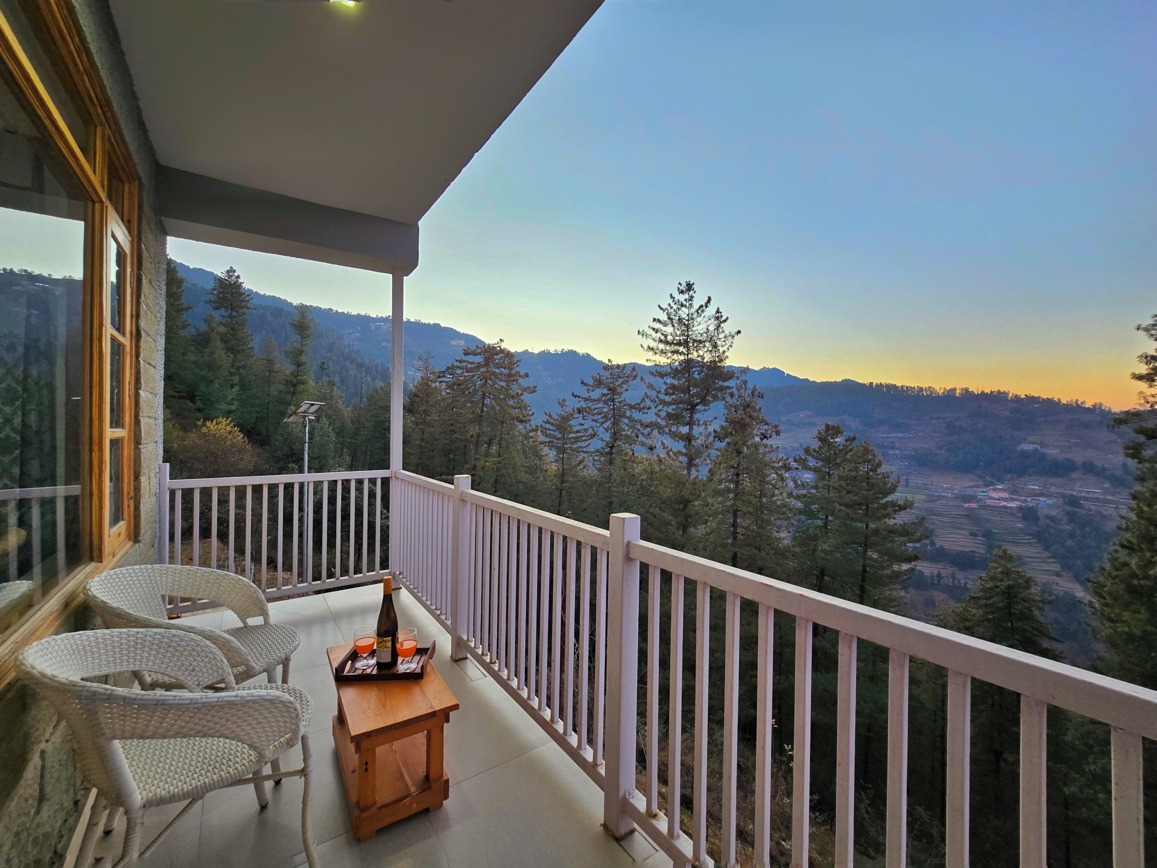 A balcony with wicker chairs, a small wooden table with a bottle and glasses, and a scenic mountain view.