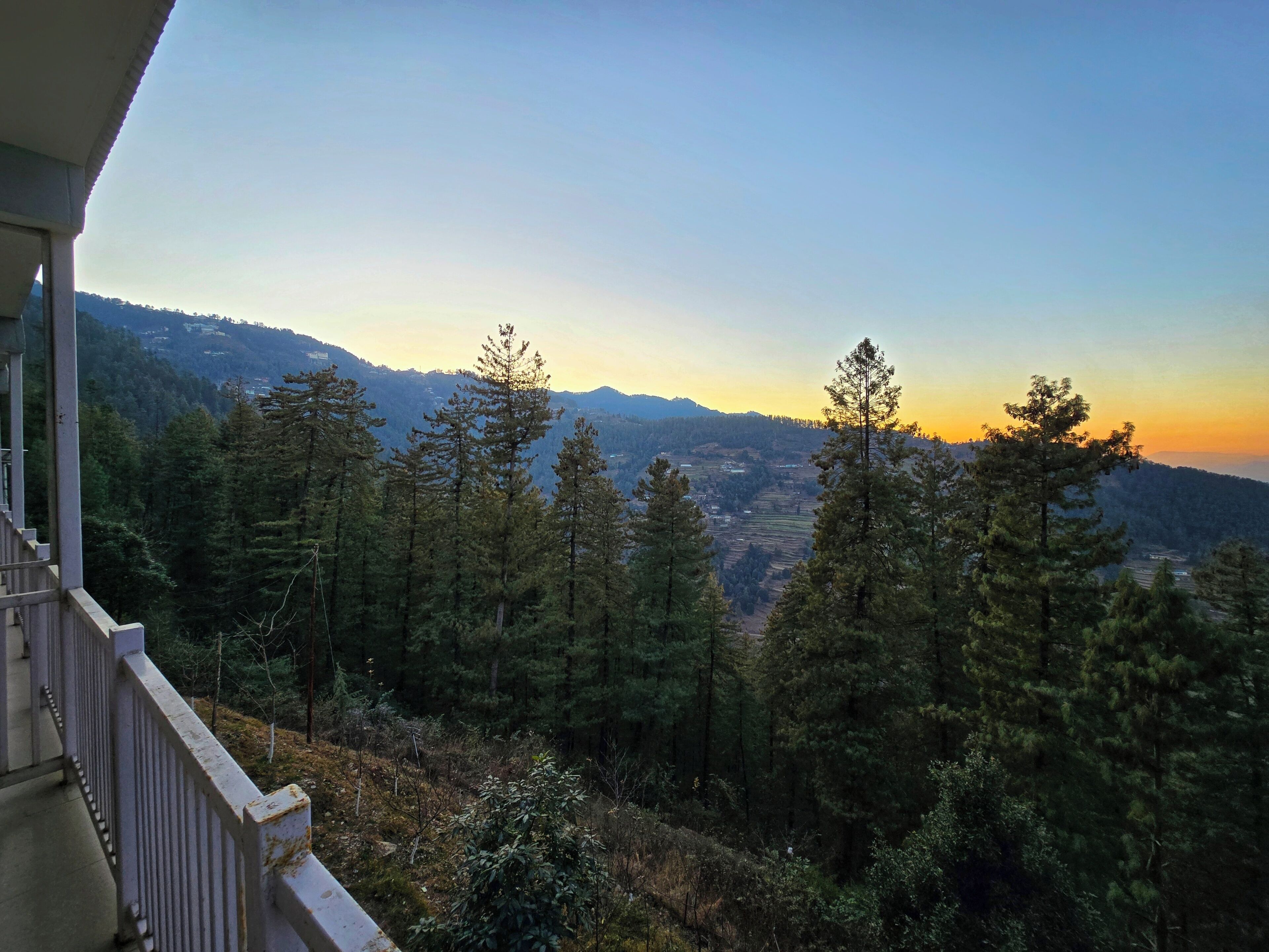En balkon med udsigt over en skovdal og fjerne bjerge i solnedgang.