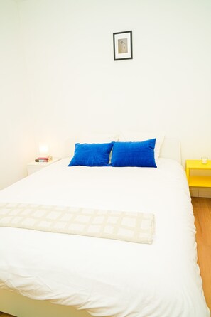 A bed with white bedding and two blue pillows, a small bedside table with a book and a candle, and a framed picture on the wall. - 1BR Apt With Living Room Near CityArea (Ghent)
