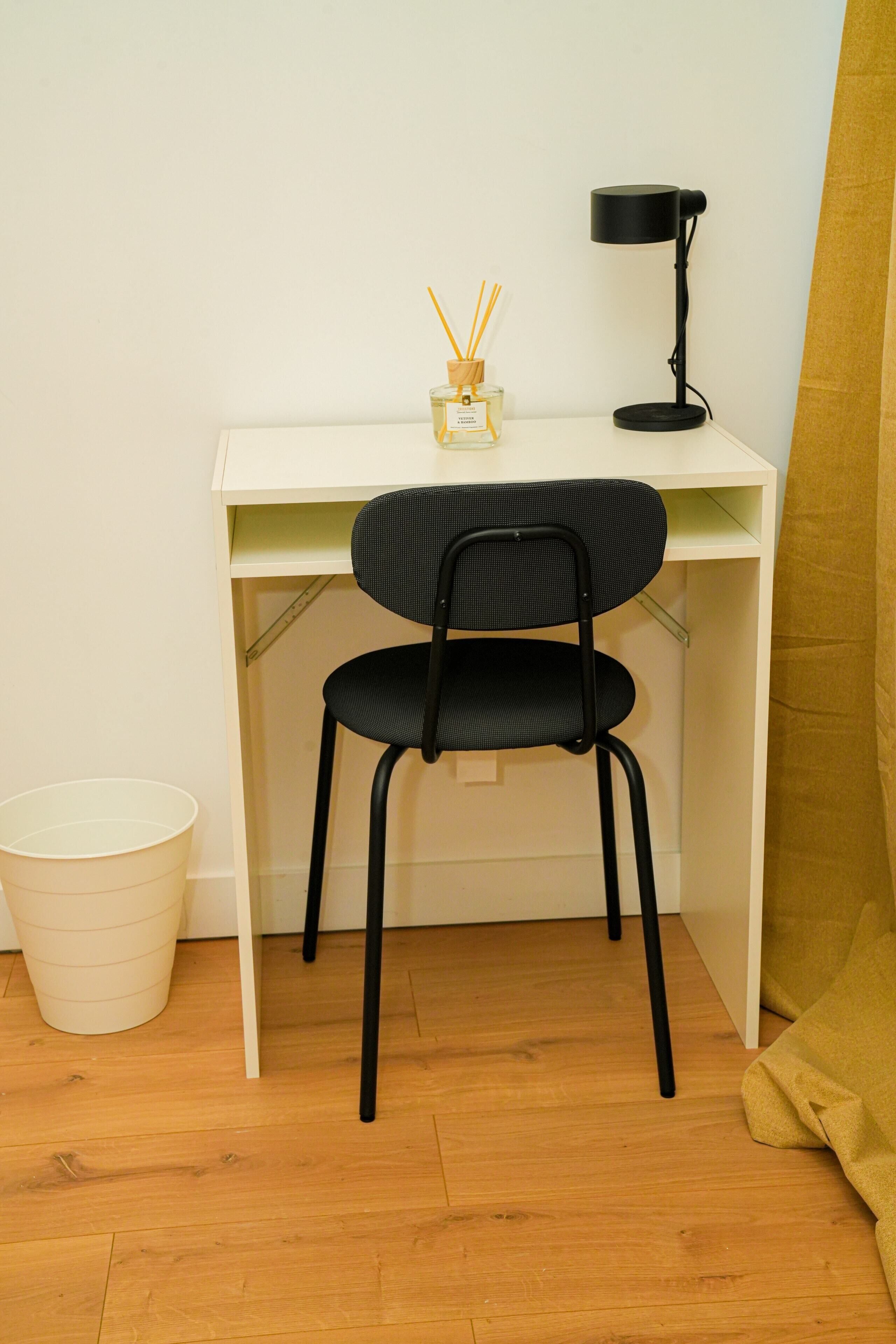 A small, white desk with a black chair, a black desk lamp, and a white trash bin.