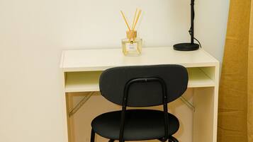 A small, white desk with a black chair, a black desk lamp, and a white trash bin.
