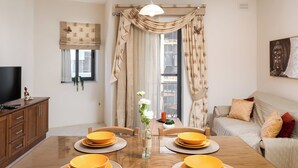 A dining area with a wooden table set for four, a television on a cabinet, and a sofa with cushions. - Spacious & Cosy APT - Msida Uni Heights (Msida)