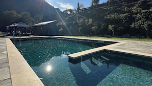 Una piscina cristalina con borde de piedra, rodeada de una zona ajardinada con árboles y una montaña al fondo.