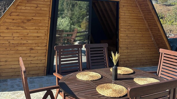 Un edificio de madera triangular con un amplio ventanal y una puerta, asientos exteriores con individuales tejidos, y vistas a árboles y colinas.