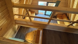 Un interior de madera con una escalera que baja a un nivel inferior, una ventana con un cartel que dice 'Welcome' y una lámpara colgante.
