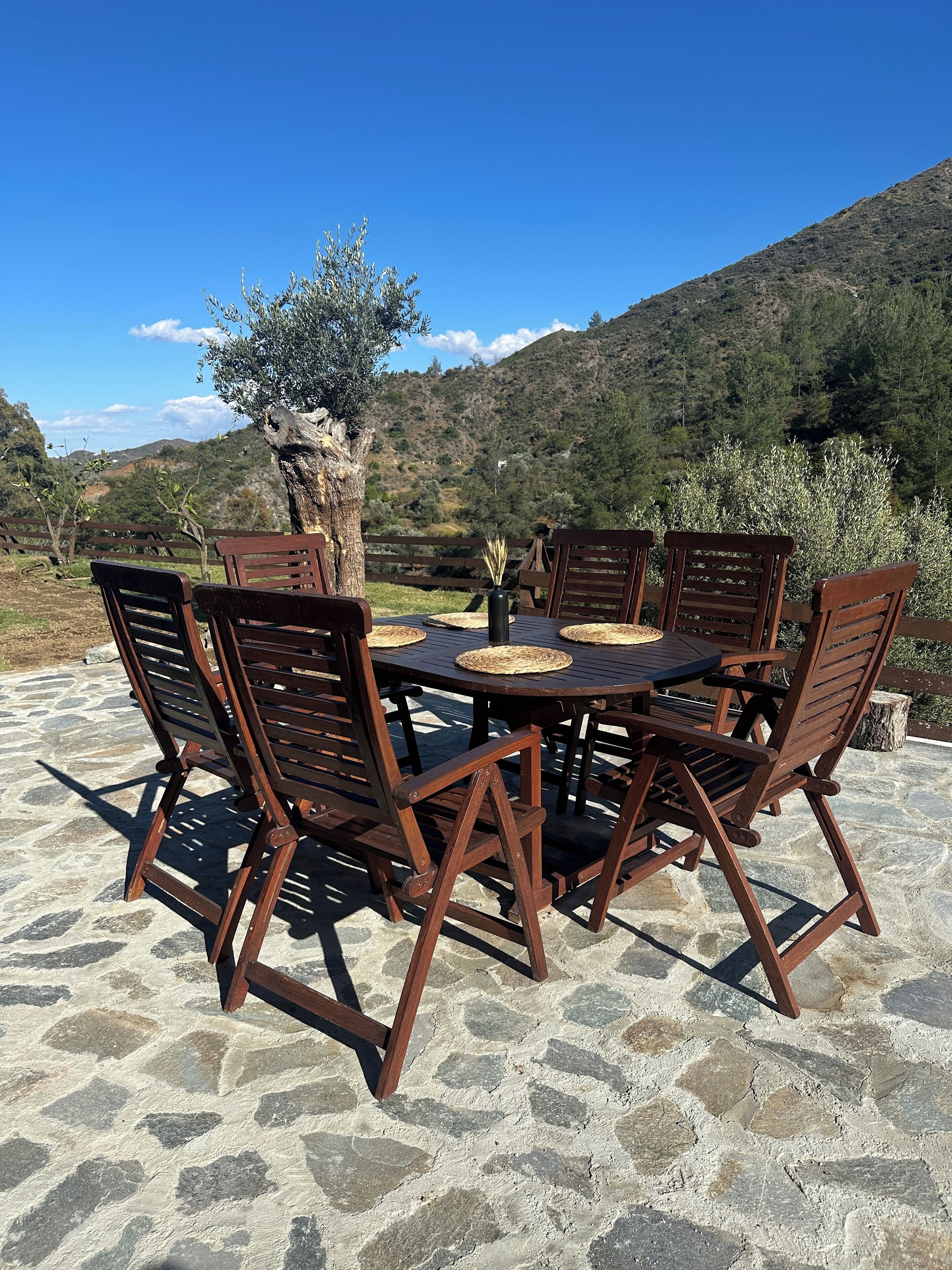 A round wooden table with six chairs, set on a stone patio with a view of a mountainous landscape.