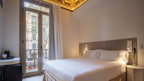 Ein Hotelzimmer mit einem großen Bett, einem Nachttisch mit einer Vase, einem Schreibtisch mit einer Kaffeemaschine und Blick auf die Stadt durch eine Balkontür.