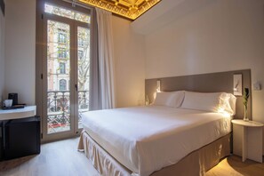 A hotel room with a large bed, a bedside table with a vase, a desk with a coffee machine, and a view of the city through a balcony door. - STORNI Barcelona (Barcelona)
