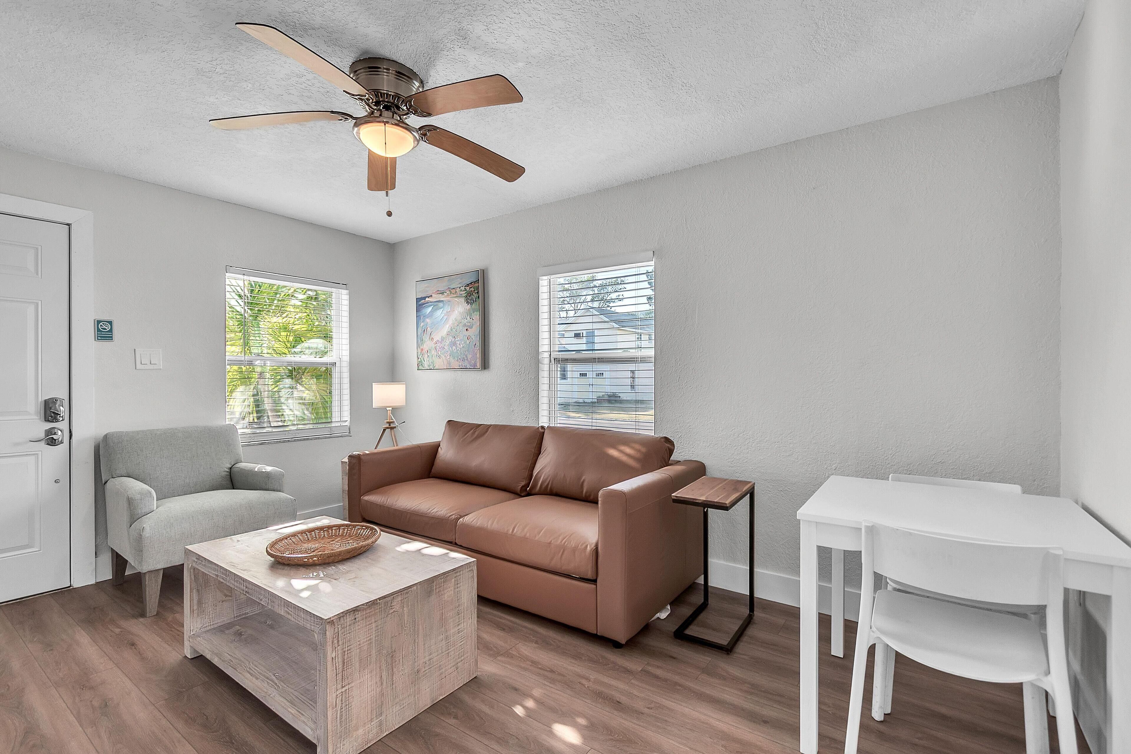 A living room with a brown leather sofa, a light grey armchair, a white table, and a ceiling fan.