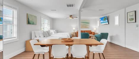 A spacious living area with a wooden dining table, white chairs, a sofa, and a ceiling fan.