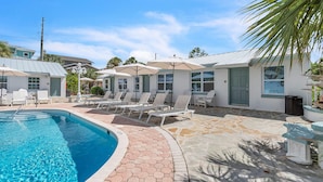 Een zwembad met ligbedden en parasols, omringd door een stenen terras en een gebouw met een groene deur.