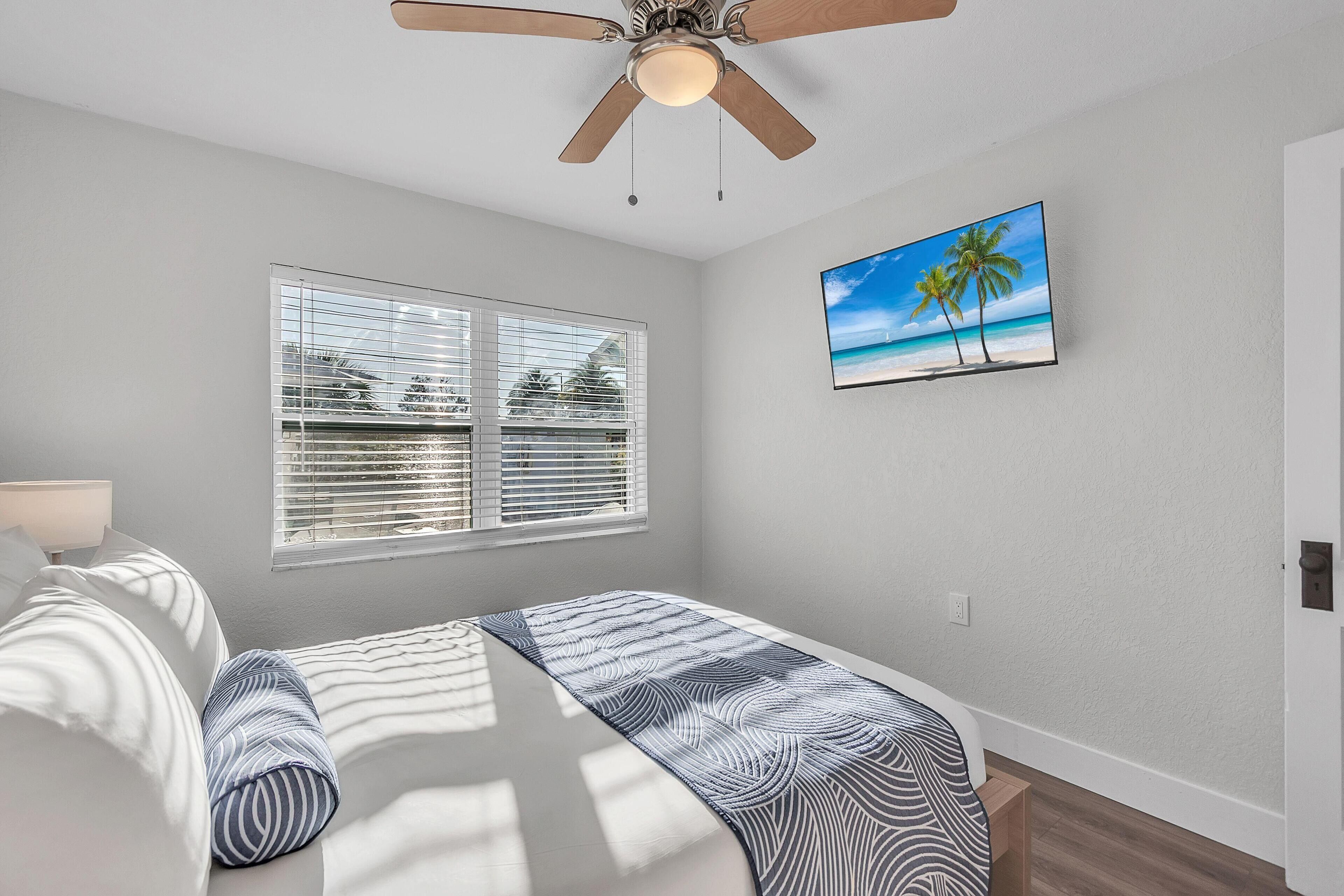 A bedroom with a bed, a ceiling fan, a window with blinds, and a painting of a beach scene.