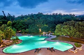 A swimming pool with a curved design, surrounded by lounge chairs and a lush, green landscape. - Tiger Camp Resort River Access (Dhikuli)