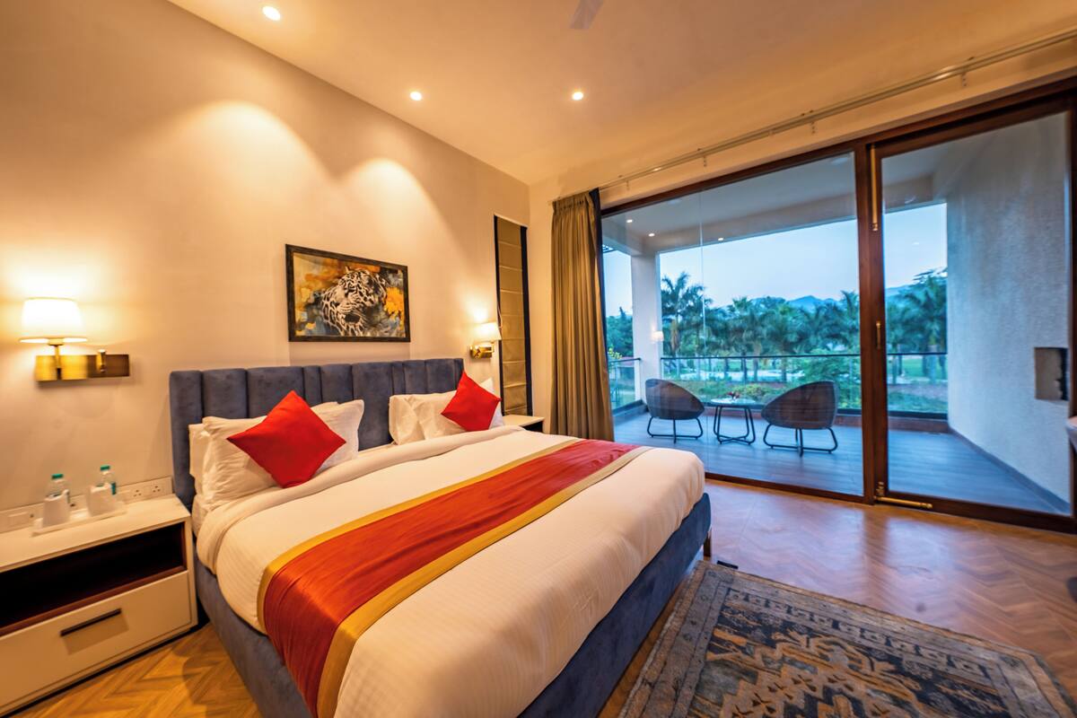 Une chambre d’hôtel moderne avec un grand lit, un tableau au mur, un balcon avec des chaises et une vue sur des palmiers.