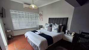 Une chambre avec un grand lit, une table de chevet, une commode, un climatiseur fixé au mur, une fenêtre avec des stores et un luminaire suspendu.