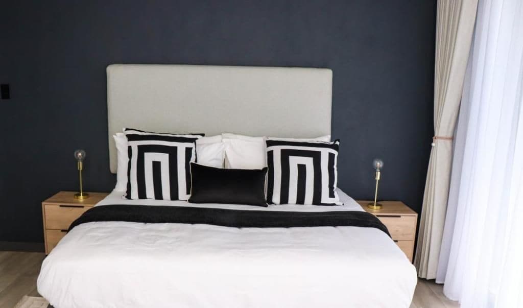 Una cama con cabecera blanca, almohadas a rayas blancas y negras y una colcha blanca. Hay dos mesitas de noche con lámparas a cada lado de la cama.