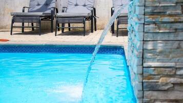 A swimming pool with clear blue water, surrounded by stone walls and grey lounge chairs.