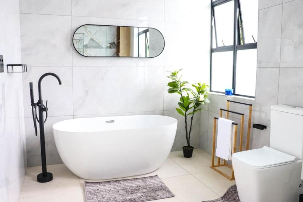 A modern bathroom with a freestanding bathtub, a large mirror, a potted plant, and a window.