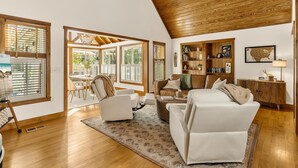 Amplia sala de estar con techo alto, vigas de madera y ventanas grandes que permiten la entrada de luz natural. Cuenta con una zona de estar con sofá y sillones, además de un comedor con mesa y sillas.
