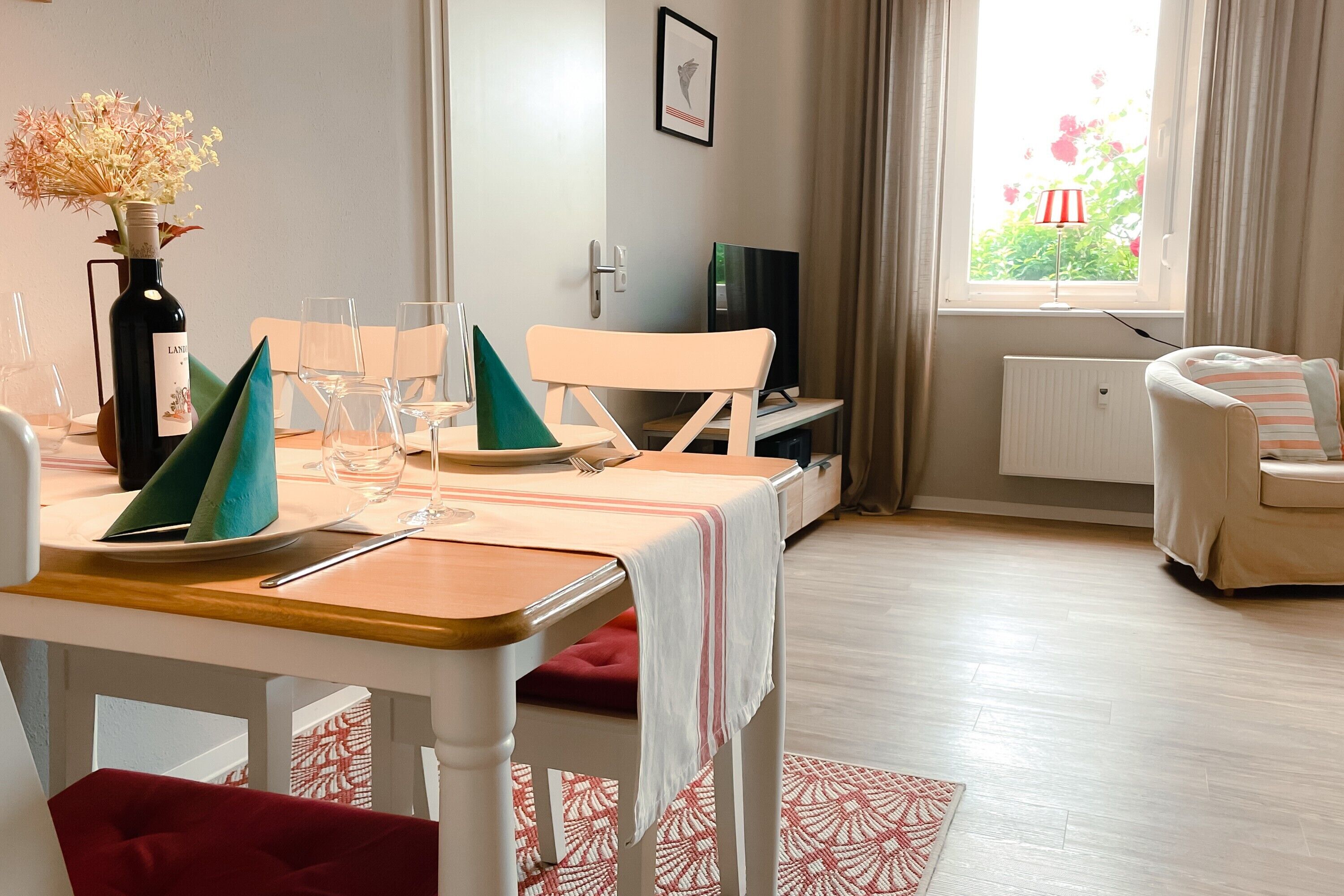 A dining area with a table set for two, a vase with flowers, a television, and a window with curtains.