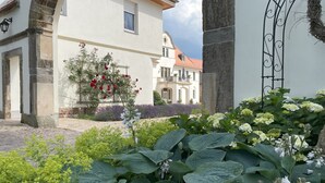 Une cour avec un bâtiment blanc, un arbre aux fleurs rouges et diverses plantes vertes.