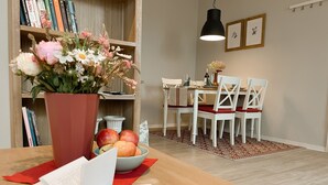Un coin repas comprenant une table, des chaises, un vase de fleurs, un saladier de fruits et une bibliothèque remplie de livres.