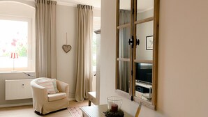Une pièce avec un radiateur blanc, un fauteuil beige orné d’un coussin à rayures, une petite table en bois sur laquelle est posée une bougie, et un grand miroir accroché au mur.