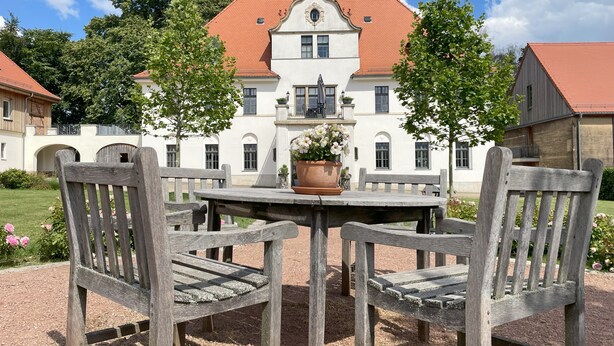 Un grand bâtiment blanc, surmonté d’un toit rouge, entouré de verdure, avec une table en bois et des chaises.