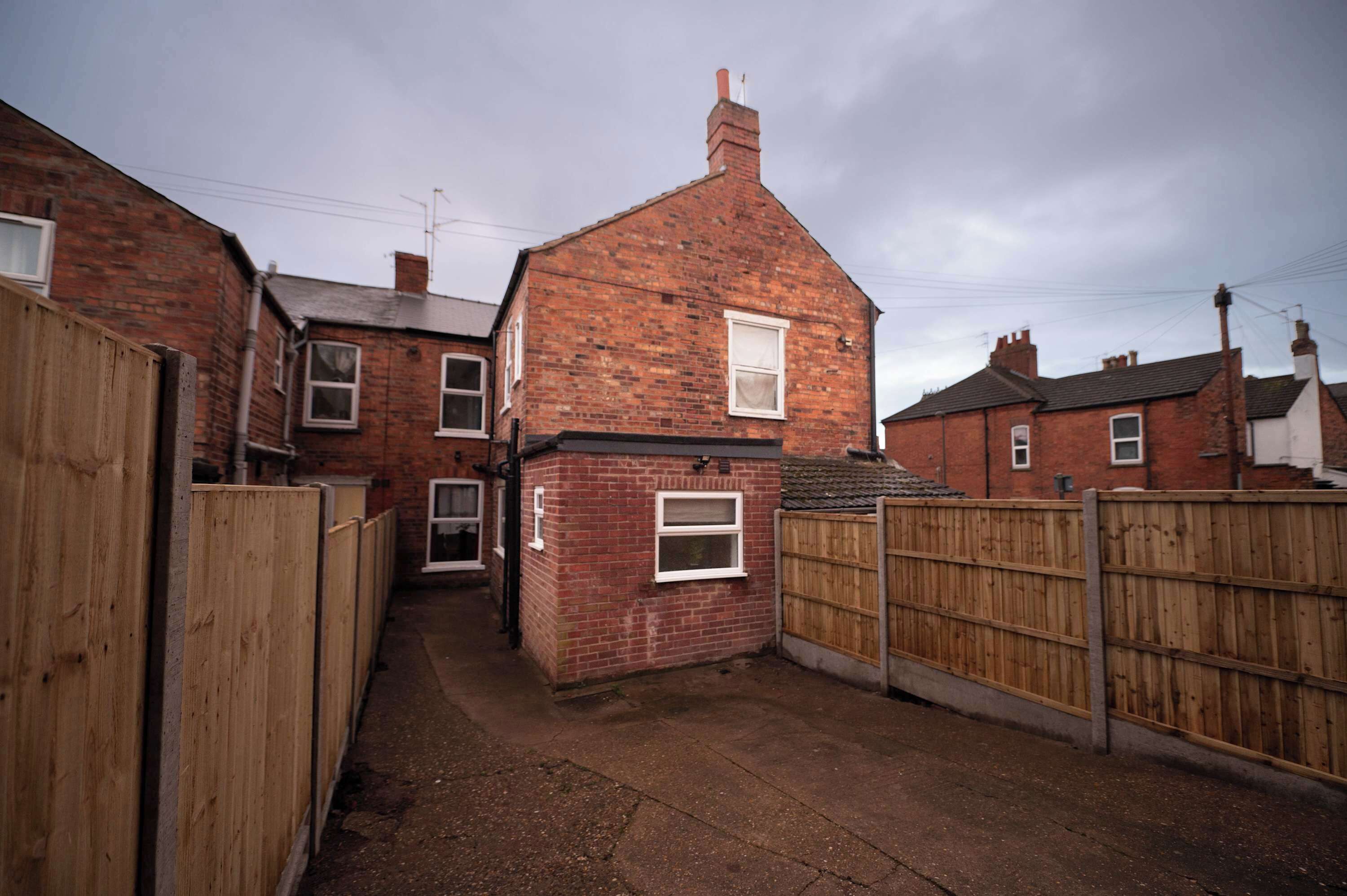 A narrow alleyway between two brick houses with a wooden fence on one side and a wooden gate on the other.