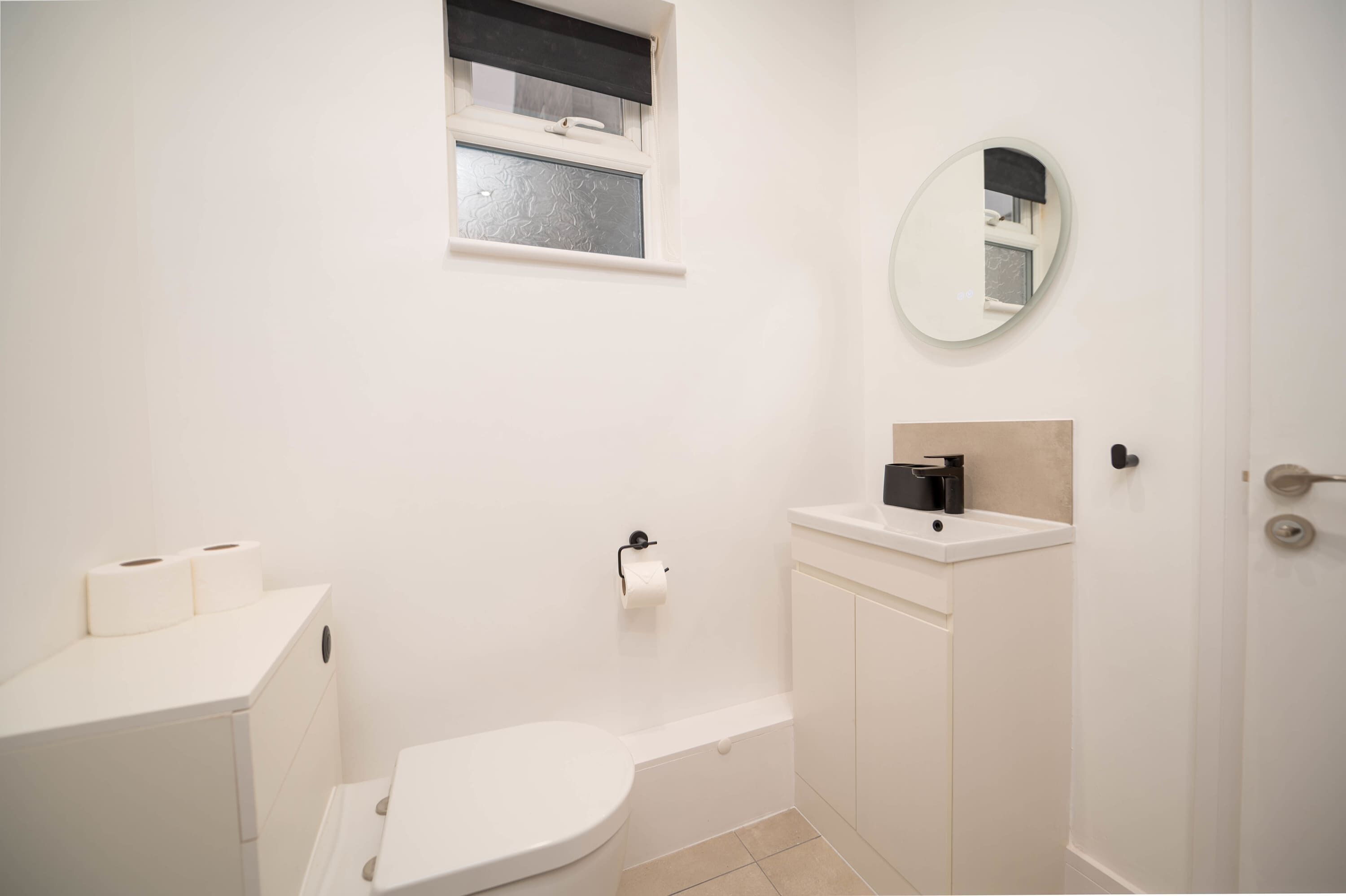 Et lille, hvidt badeværelse med toilet, vask, spejl og et vindue.