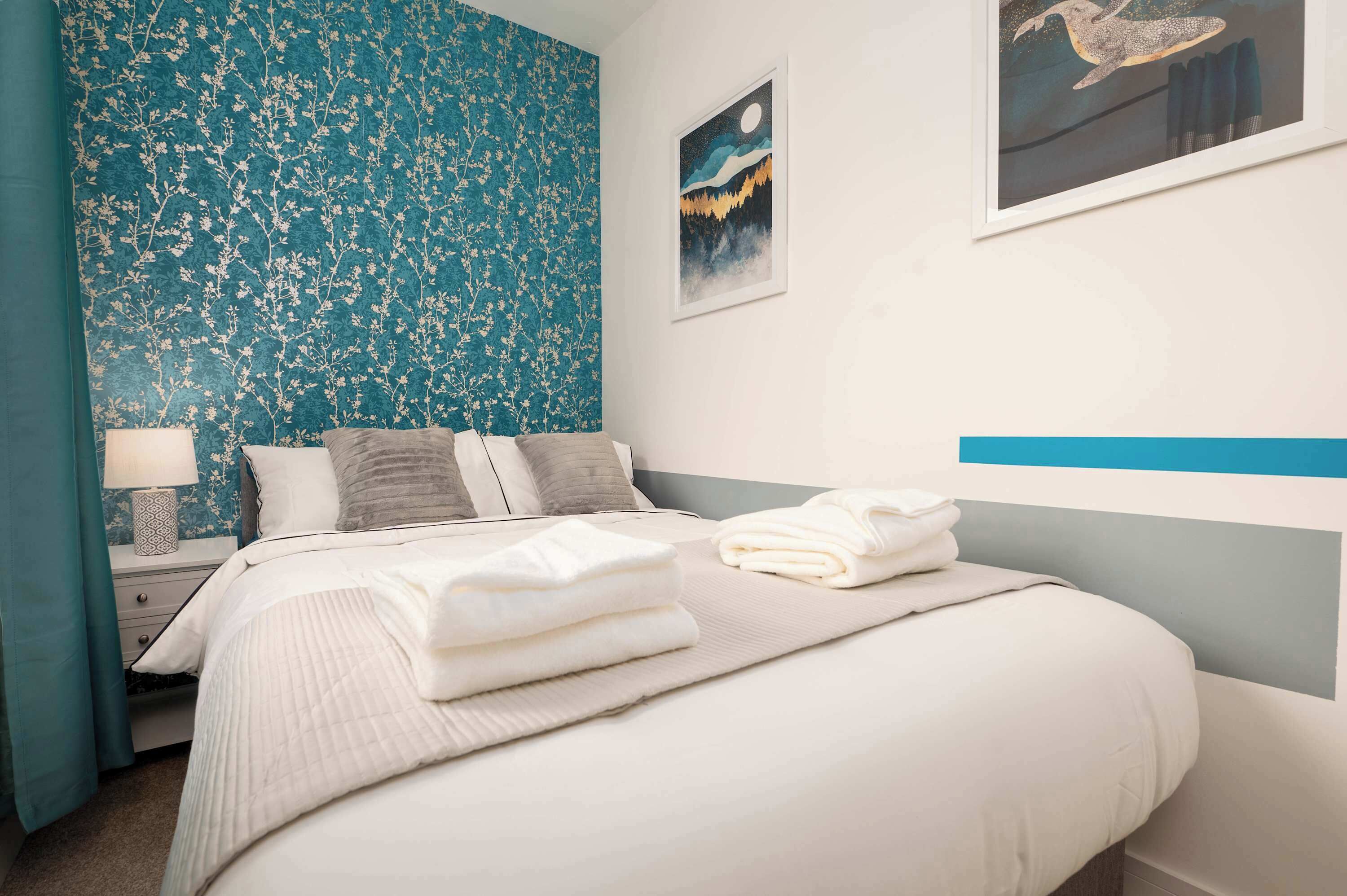 A neatly made bed with white linens and pillows, a bedside table with a lamp, and a wall with a floral pattern and artwork.