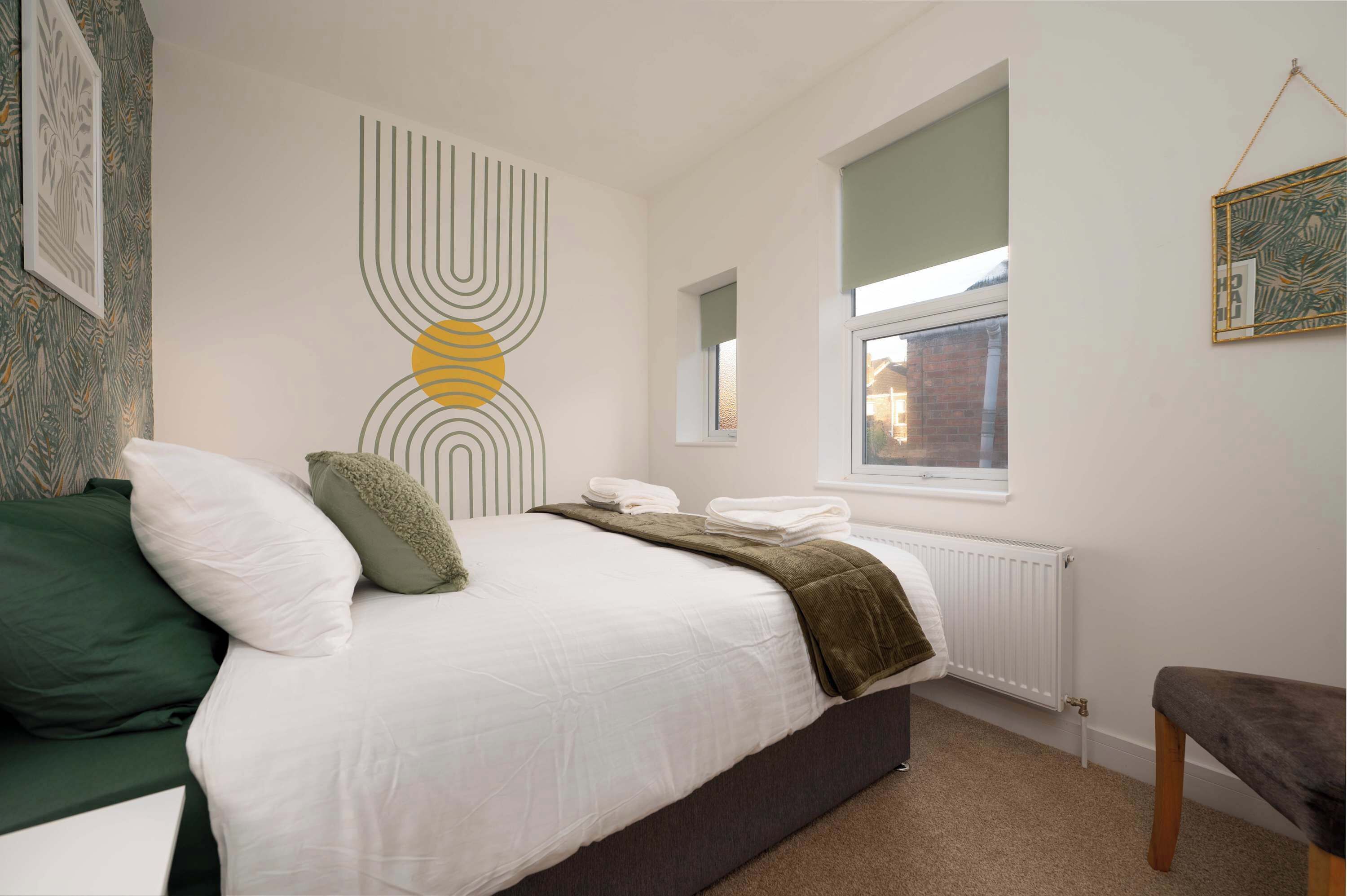 Un dormitorio con cama, almohadas, radiador, ventana y obras de arte en las paredes.