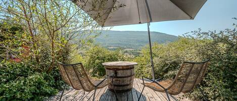 Una terraza de madera con dos sillas de mimbre y una mesa redonda de madera, una sombrilla y vistas a un paisaje montañoso.