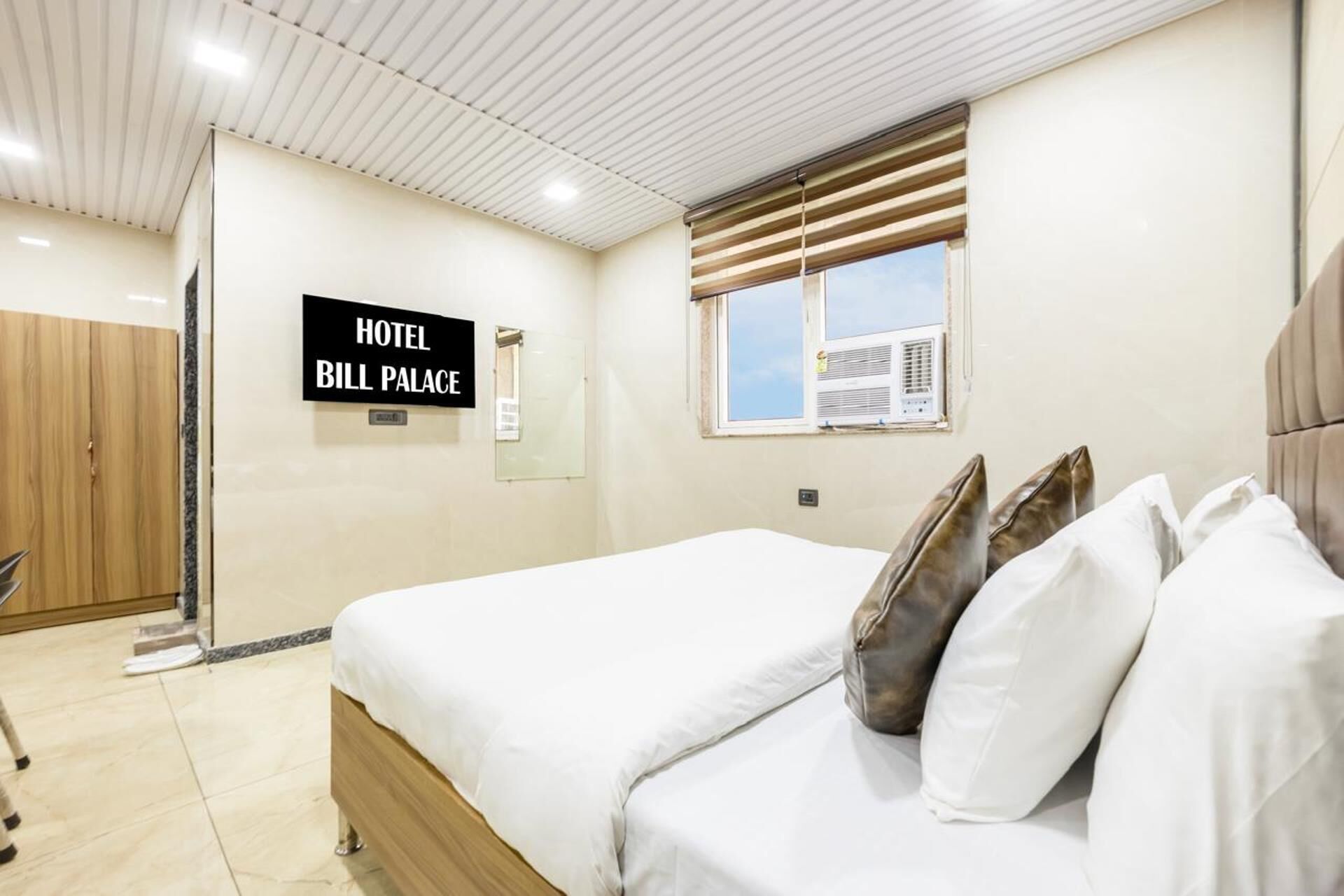 Una habitación de hotel con una cama, almohadas, una ventana con persianas y un techo con iluminación empotrada, y un letrero que dice 'HOTEL BILL PALACE'.