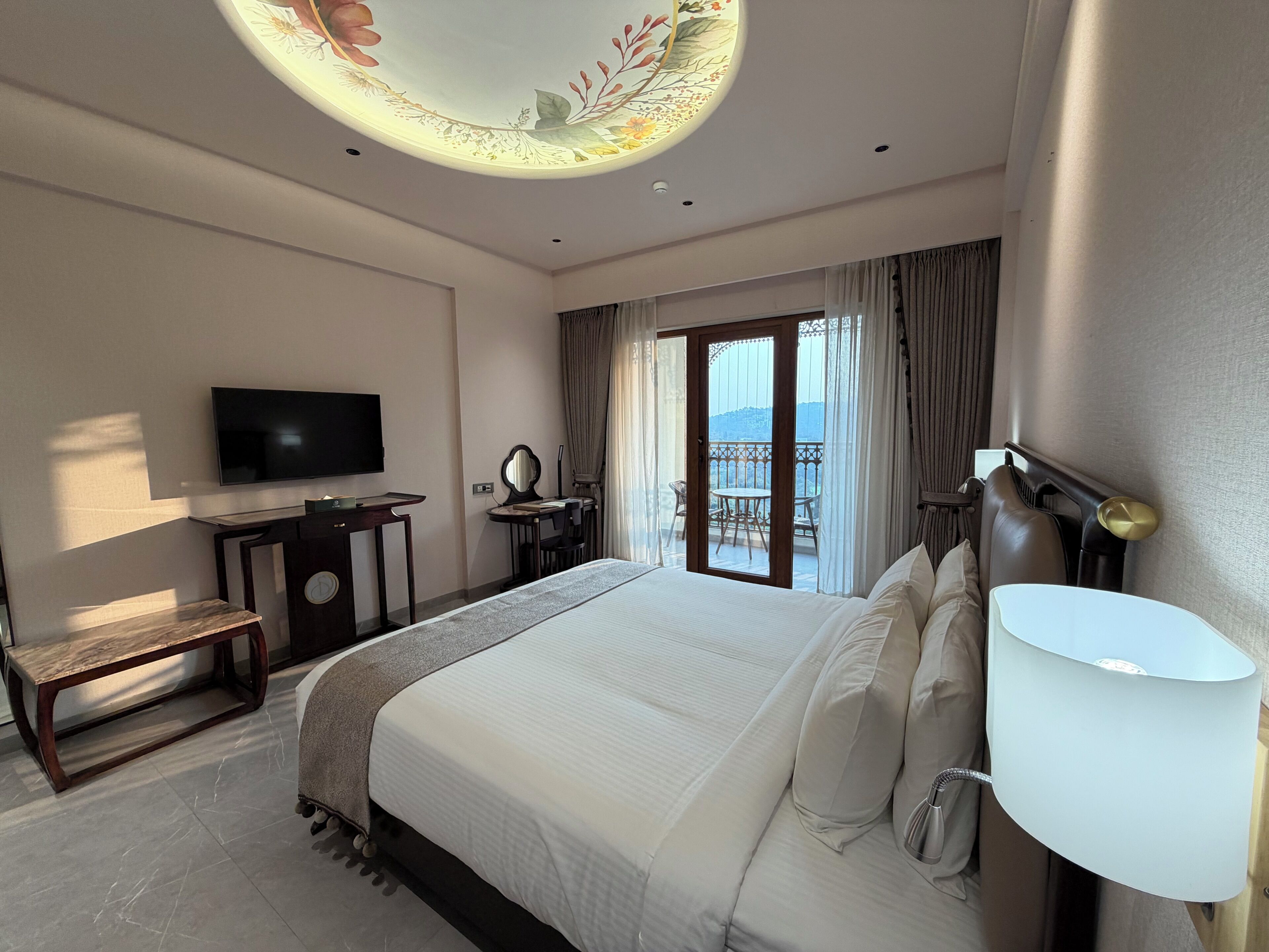 Habitación de hotel con cama, televisión, escritorio, silla, lámpara y vistas al exterior.