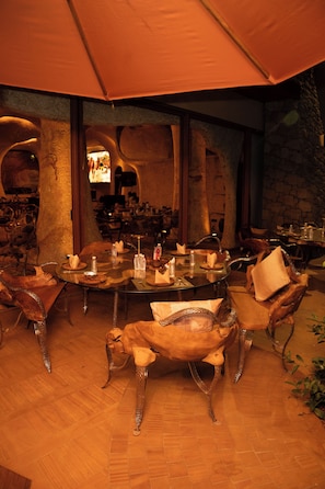 A restaurant with a rustic interior featuring stone walls and wooden furniture. There is a round table with chairs, and the room has a warm, dimly lit ambiance. - De Lush Pune (Paud)