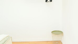 Una habitación con pared blanca, suelo de madera, cama blanca con manta verde, estante blanco con toallas y un cesto de basura pequeño.