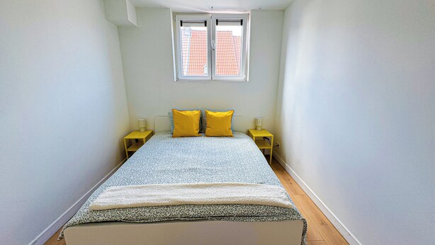 Una habitación pequeña y estrecha con una cama, dos mesitas de noche y una ventana.