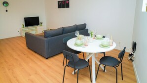 Una sala moderna con un sofá gris, una mesa de comedor blanca con cuatro sillas y un televisor de pantalla plana sobre un mueble blanco.