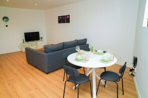A modern living room with a grey sofa, a white dining table with four chairs, and a flat-screen TV on a white stand. - Cozy 1BR With Living Room Near City (Ghent)