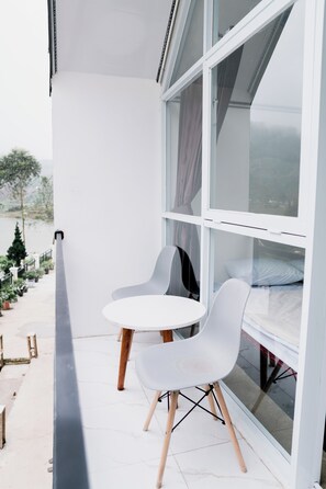 Un balcón con una mesa blanca y dos sillas blancas, una cama con ropa de cama blanca, y vistas a un lago y árboles.