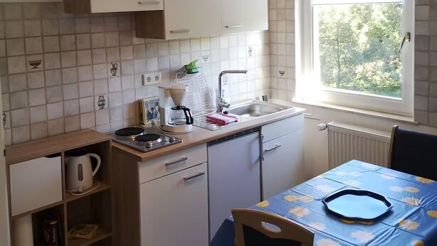 Una cocina compacta con armarios blancos, una ventana y una mesa con mantel azul.