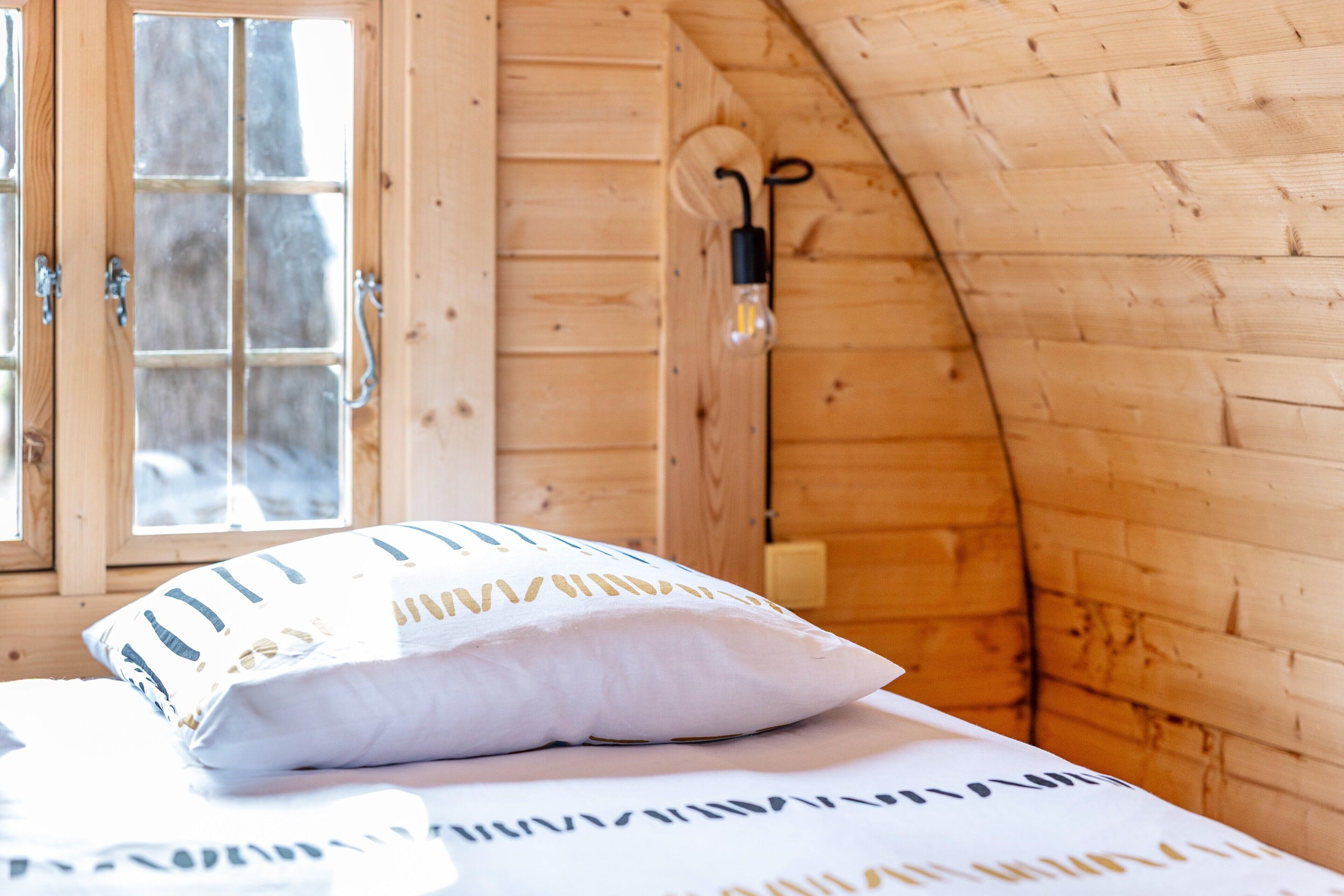 A wooden cabin room with a bed, a window, and a wall-mounted light fixture.