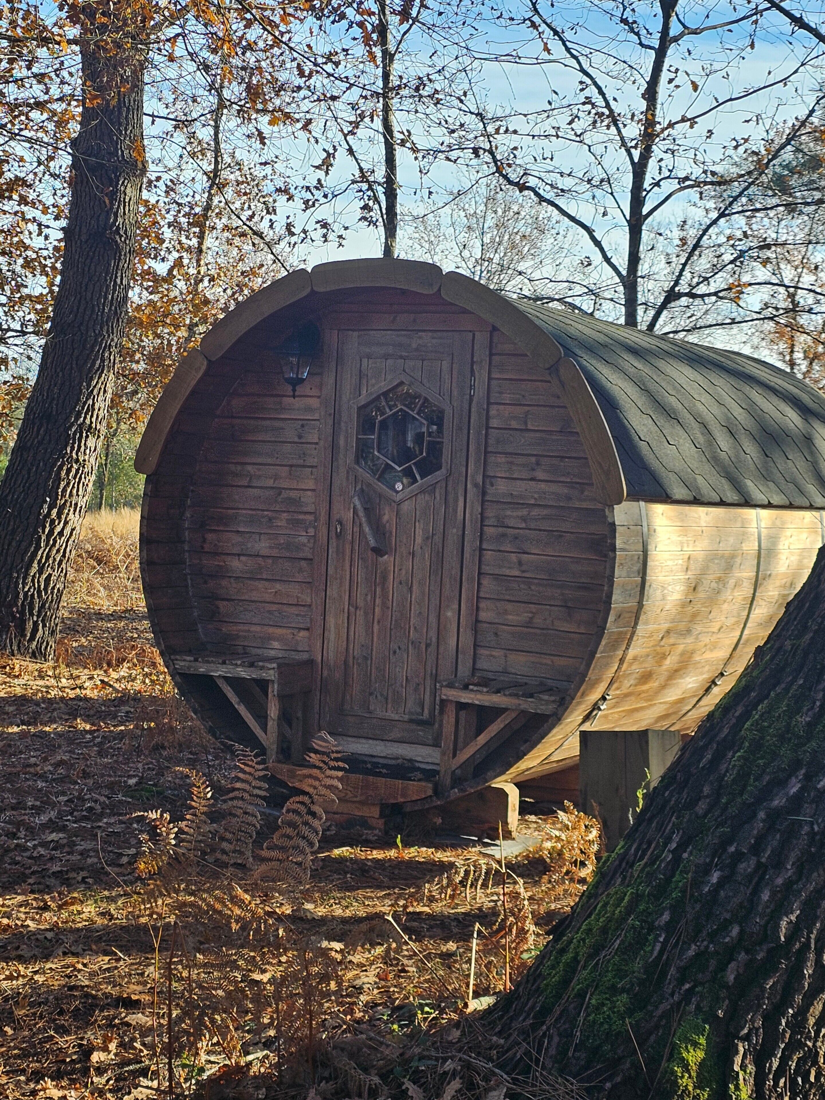 A wooden barrel-shaped structure with a hexagonal door, surrounded by trees and dry leaves.