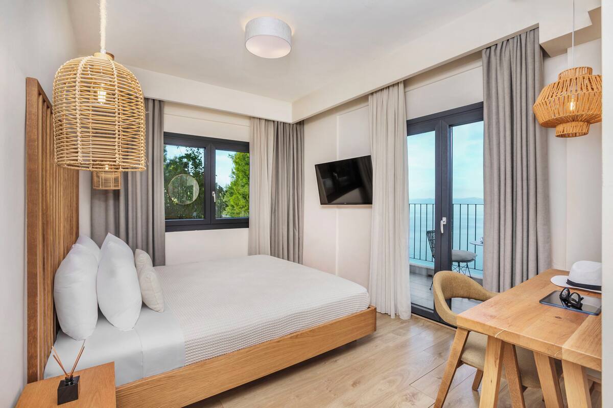 Habitación de hotel moderna con una cama grande, una mesa de comedor, un televisor y un balcón con vistas al exterior.