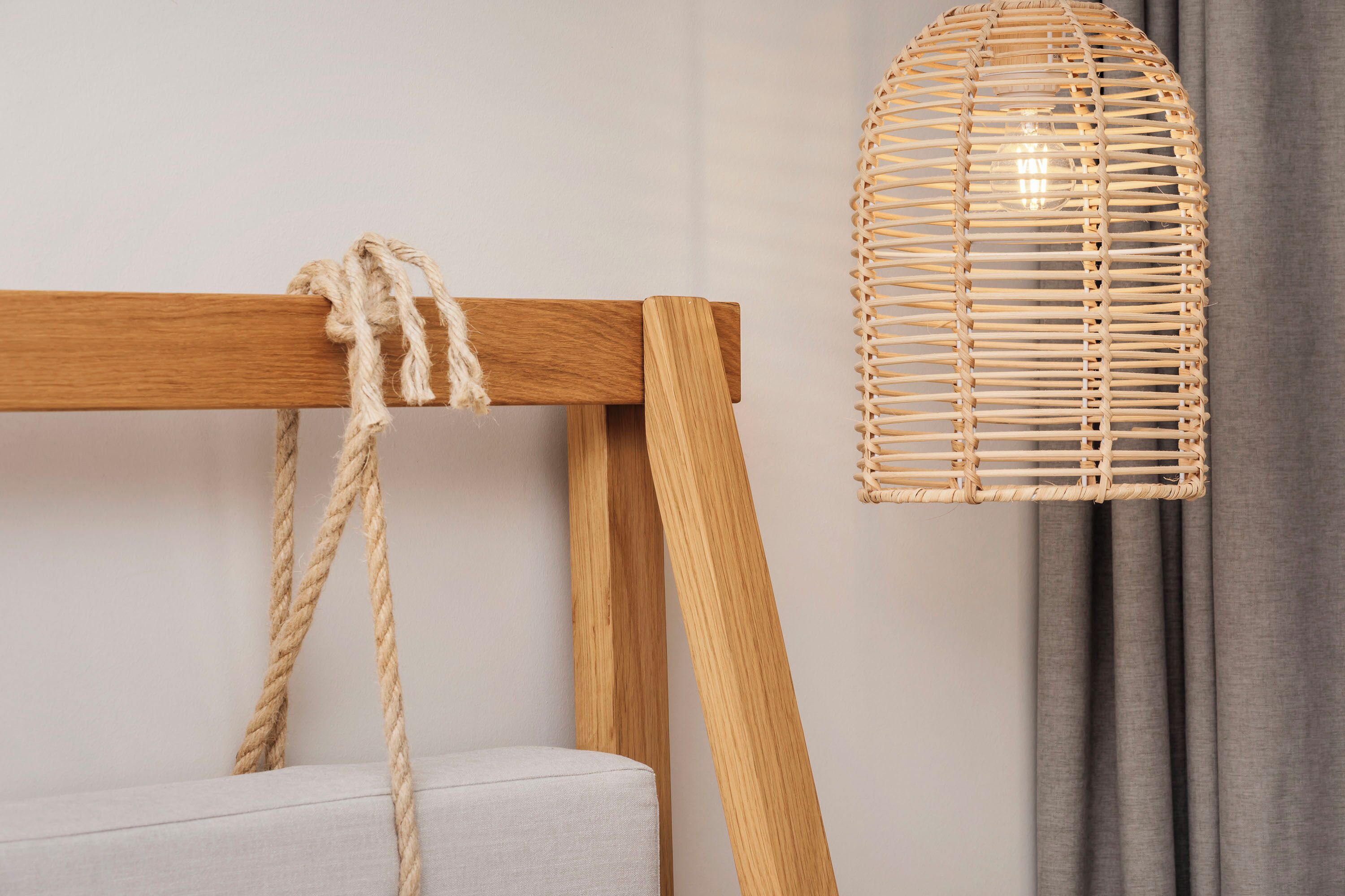 Une lampe en osier, posée sur un pied en bois auquel est attachée une corde, le tout adossé à un mur uni, avec un rideau gris en arrière-plan.