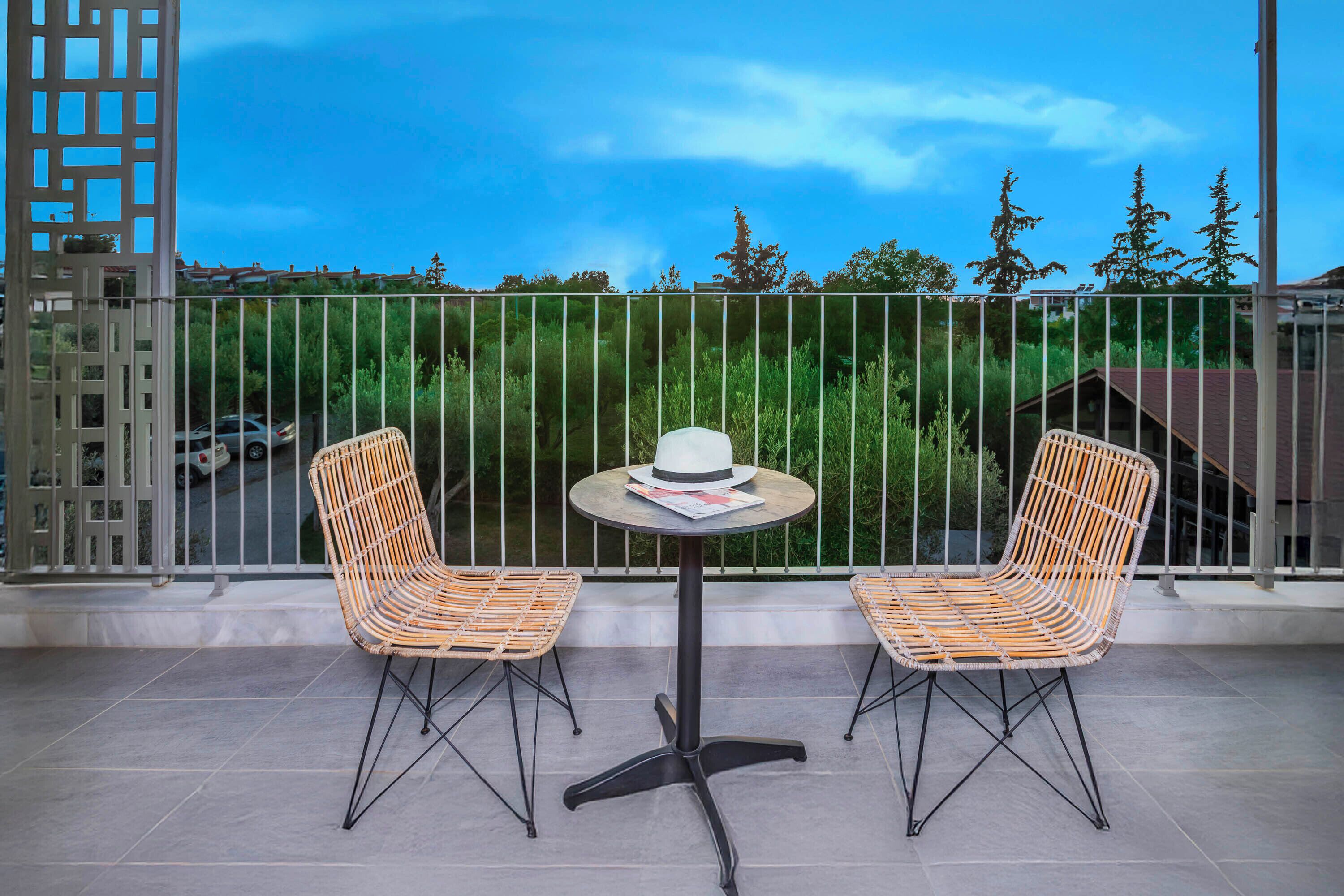 Ein Balkon mit zwei Korbstühlen und einem kleinen runden Tisch, mit Blick auf eine Grünanlage mit Bäumen und einem Gebäude im Hintergrund.